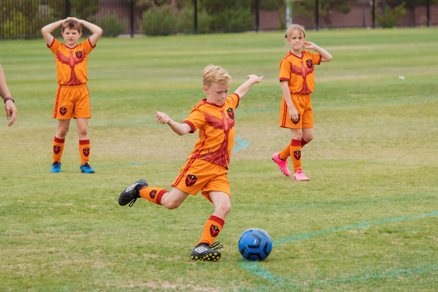 A boy in the Phoenix SC kit kicks the ball while teammates watch.